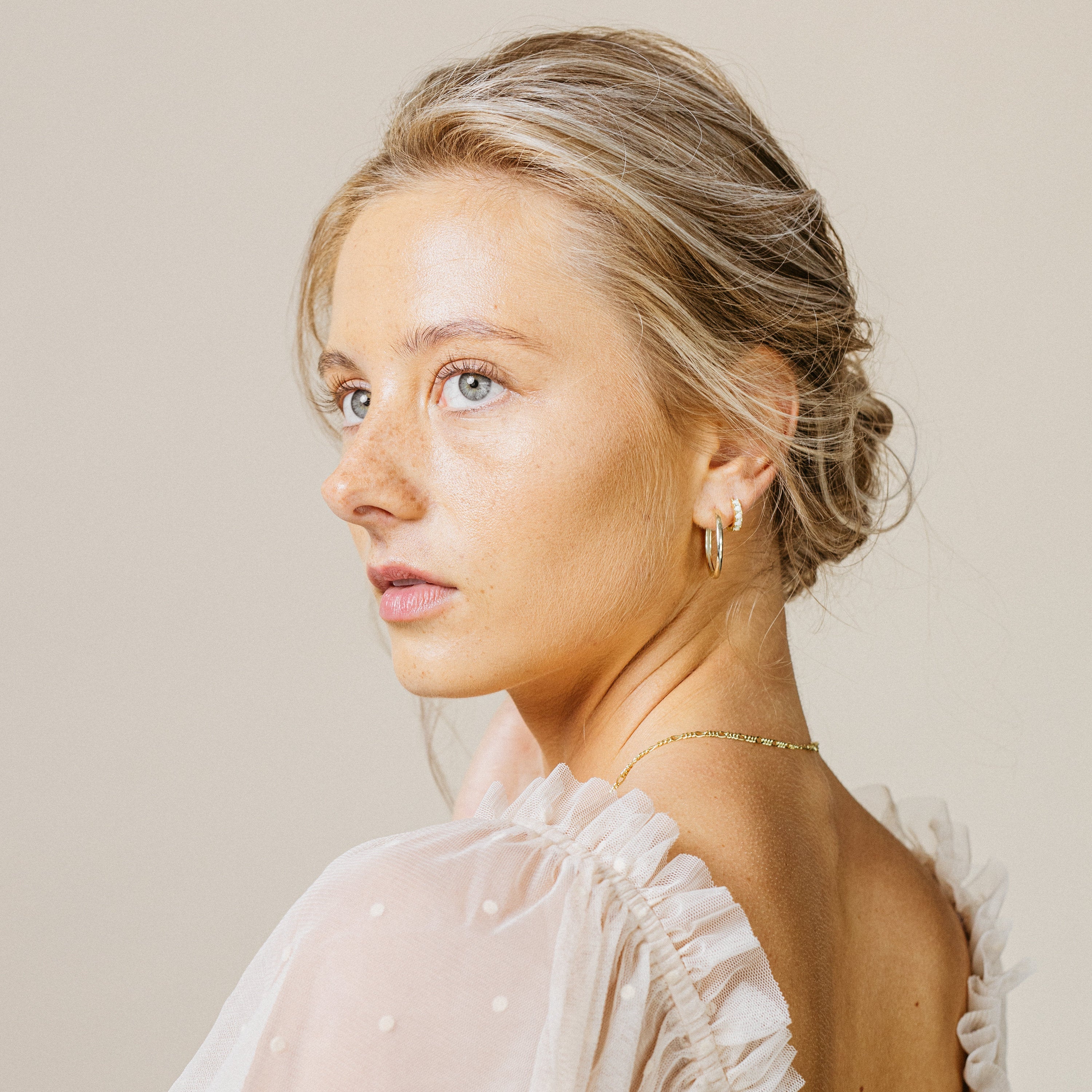 Woman with styled hair, gold hoop earrings and a light-colored top against a neutral background
