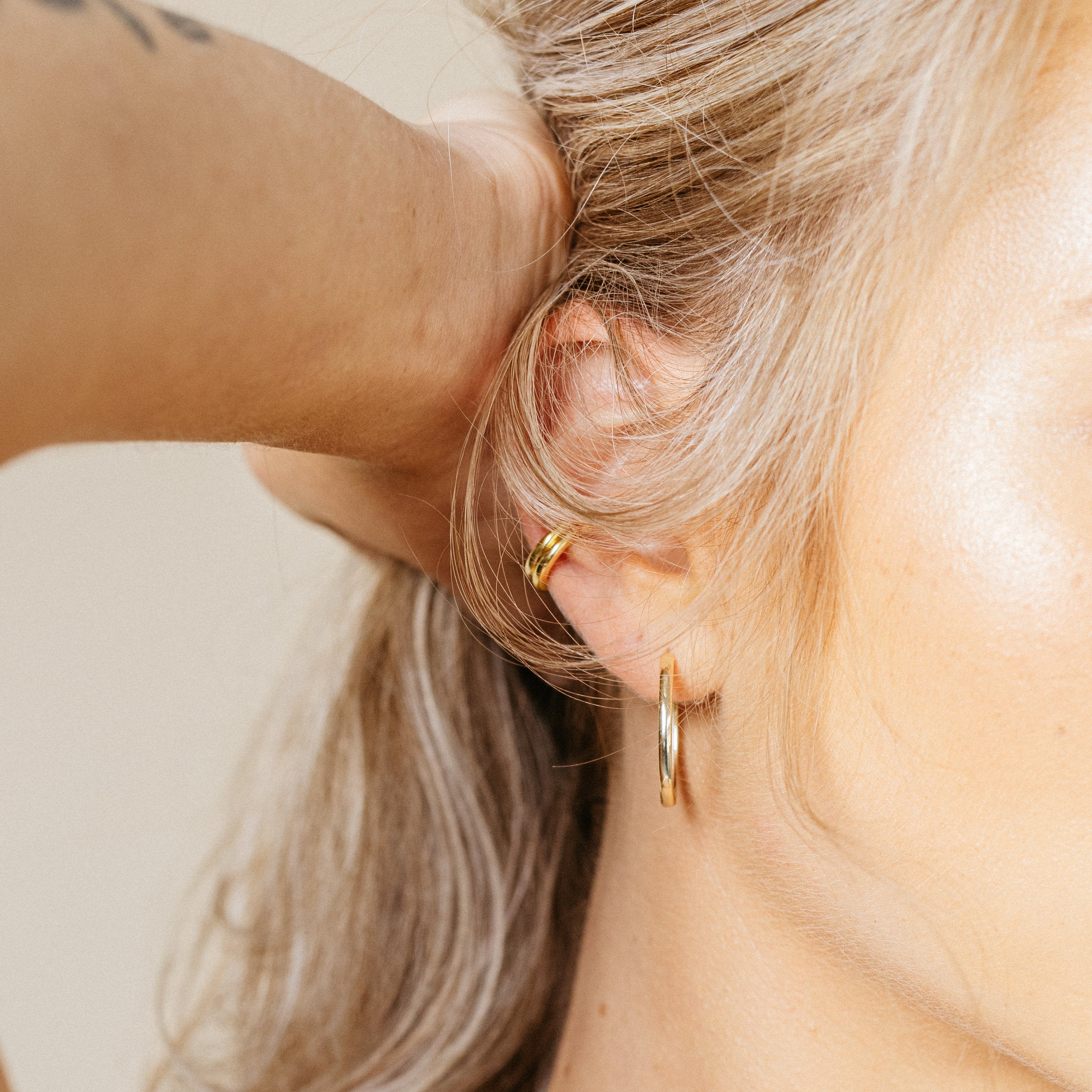 Close-up of a person wearing 2 gold cuff earrings styled with a gold hoop earring in a neutral background