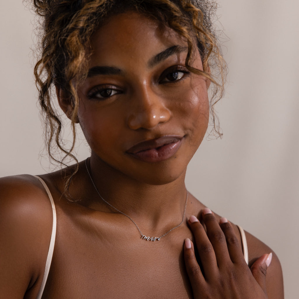 A woman with curly hair wears the Mrs. Necklace and a spaghetti strap top, smiling softly—an ideal wedding gift for any bride-to-be.
