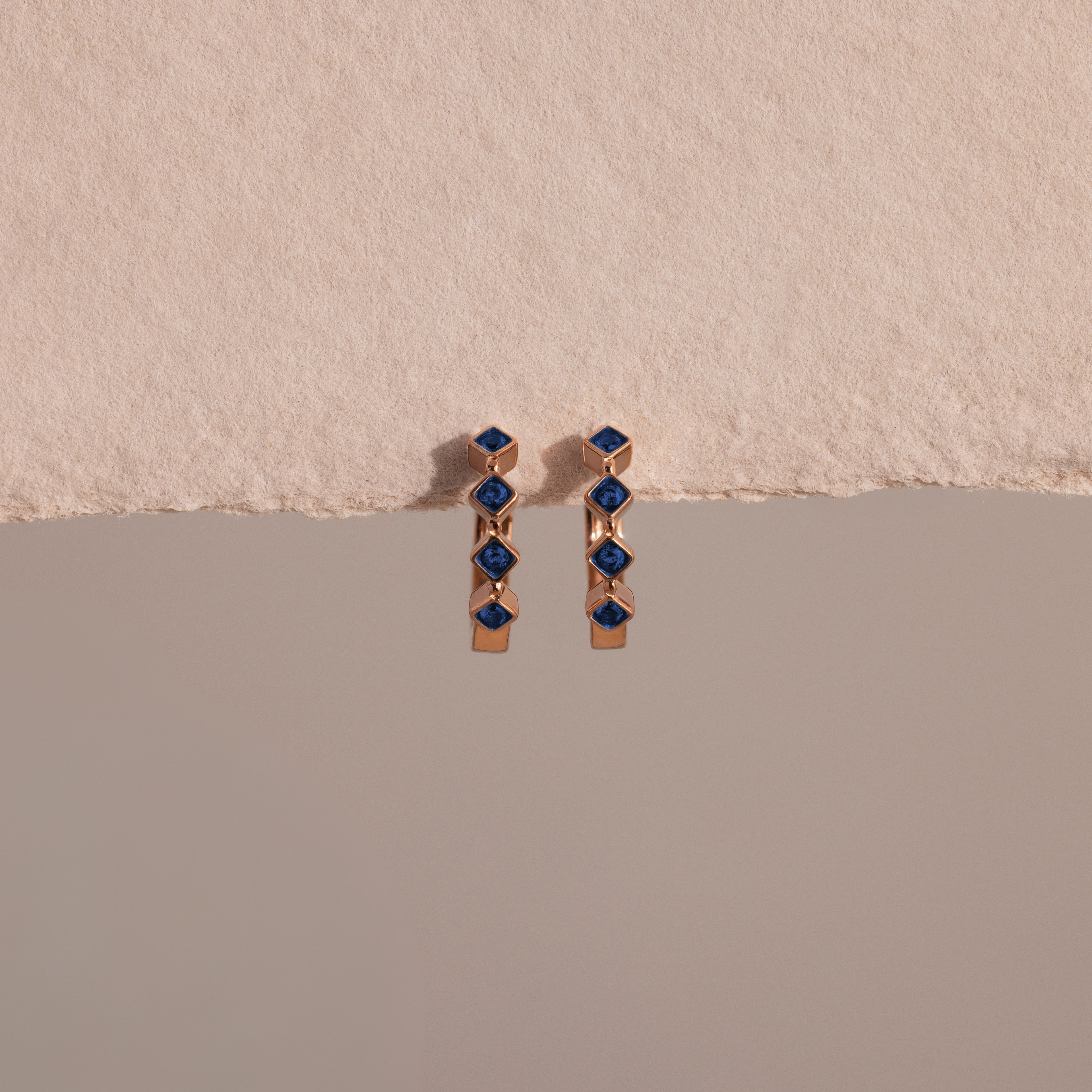 Geometric Sapphire Huggies: Gold earrings featuring a vertical row of blue sapphires, inspired by geometric birthstone designs with diamond accents, displayed on a textured beige and grey background.