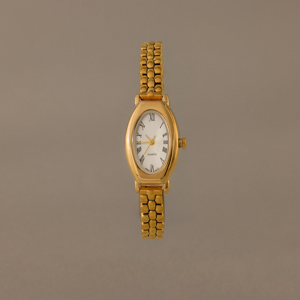 The Grid Chain Timepiece features a gold oval dial, white face, Roman numerals, and a vintage-inspired bracelet on a plain neutral background.