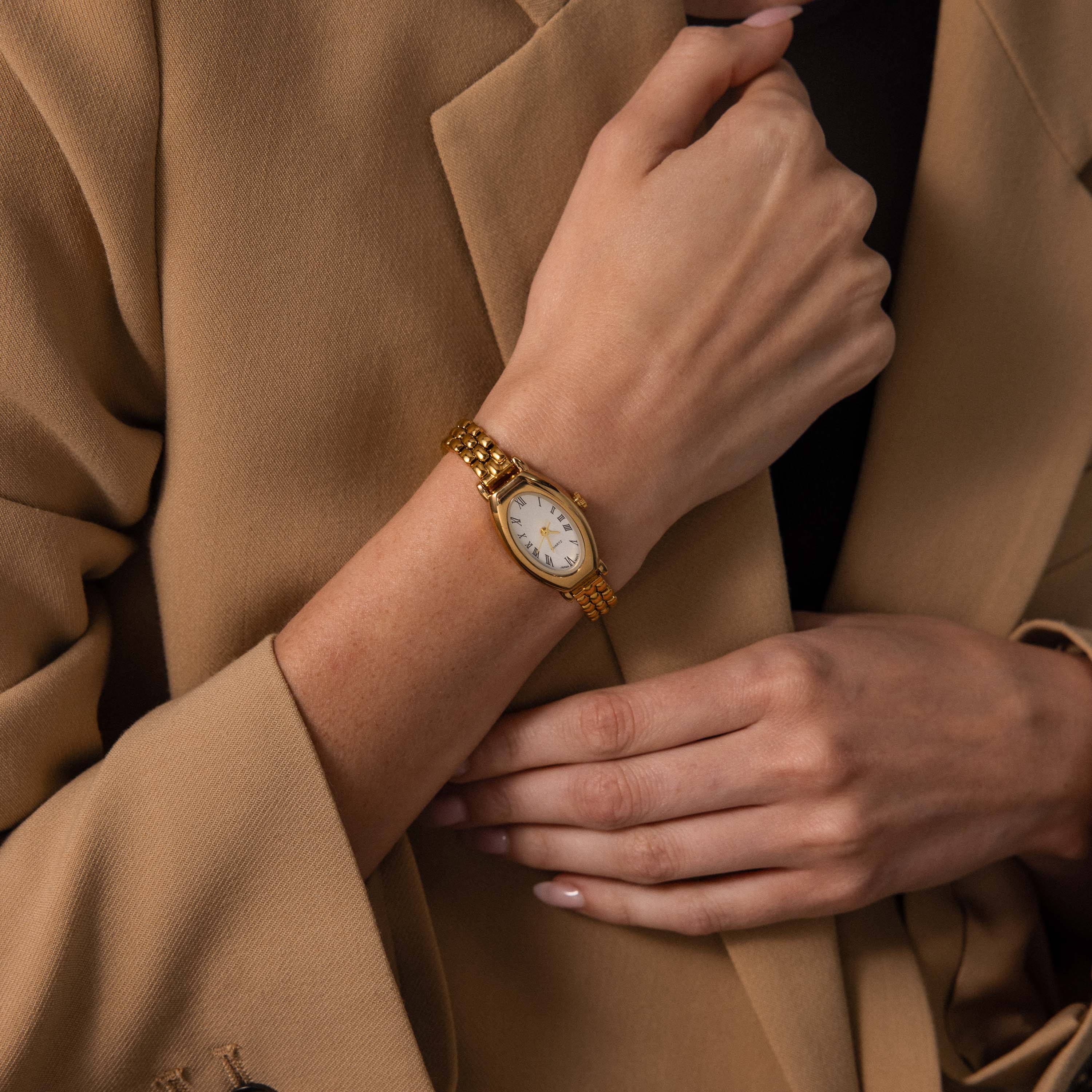 Woman wearing a beige blazer gently touches her wrist, showcasing the Grid Chain Timepiece.