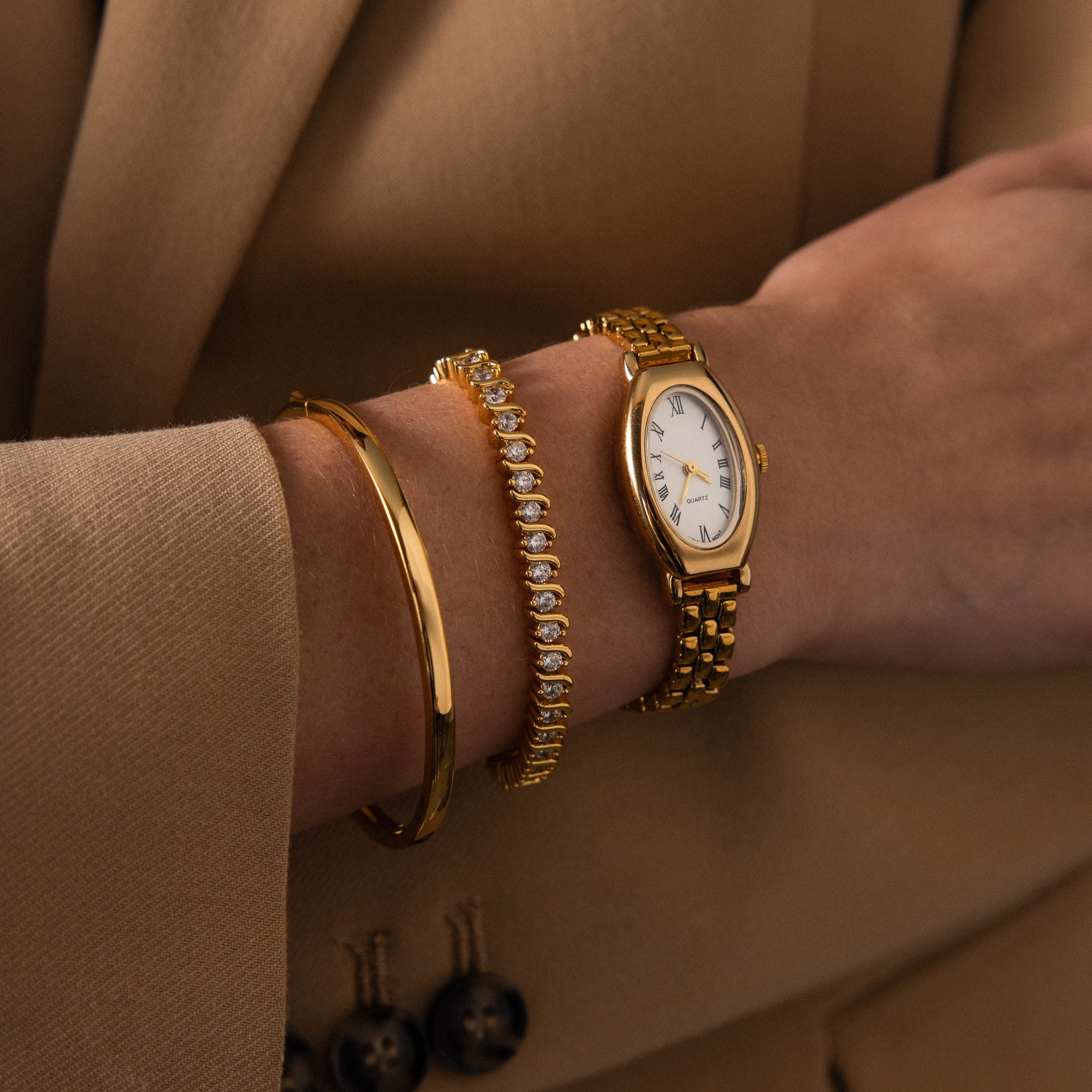 A person in a beige suit wears the Grid Chain Timepiece—a gold watch with vintage charm—paired with a diamond bracelet.