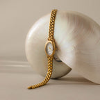 The Grid Chain Timepiece with a gold oval dial rests atop a large, iridescent seashell on a neutral background.