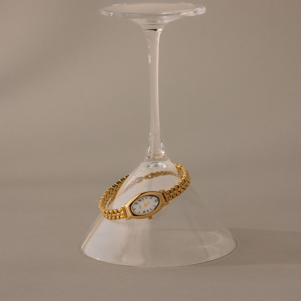 The Grid Chain Timepiece, a vintage-inspired bracelet oval dial watch, is elegantly draped around the stem of an upside-down wine glass on a neutral backdrop.