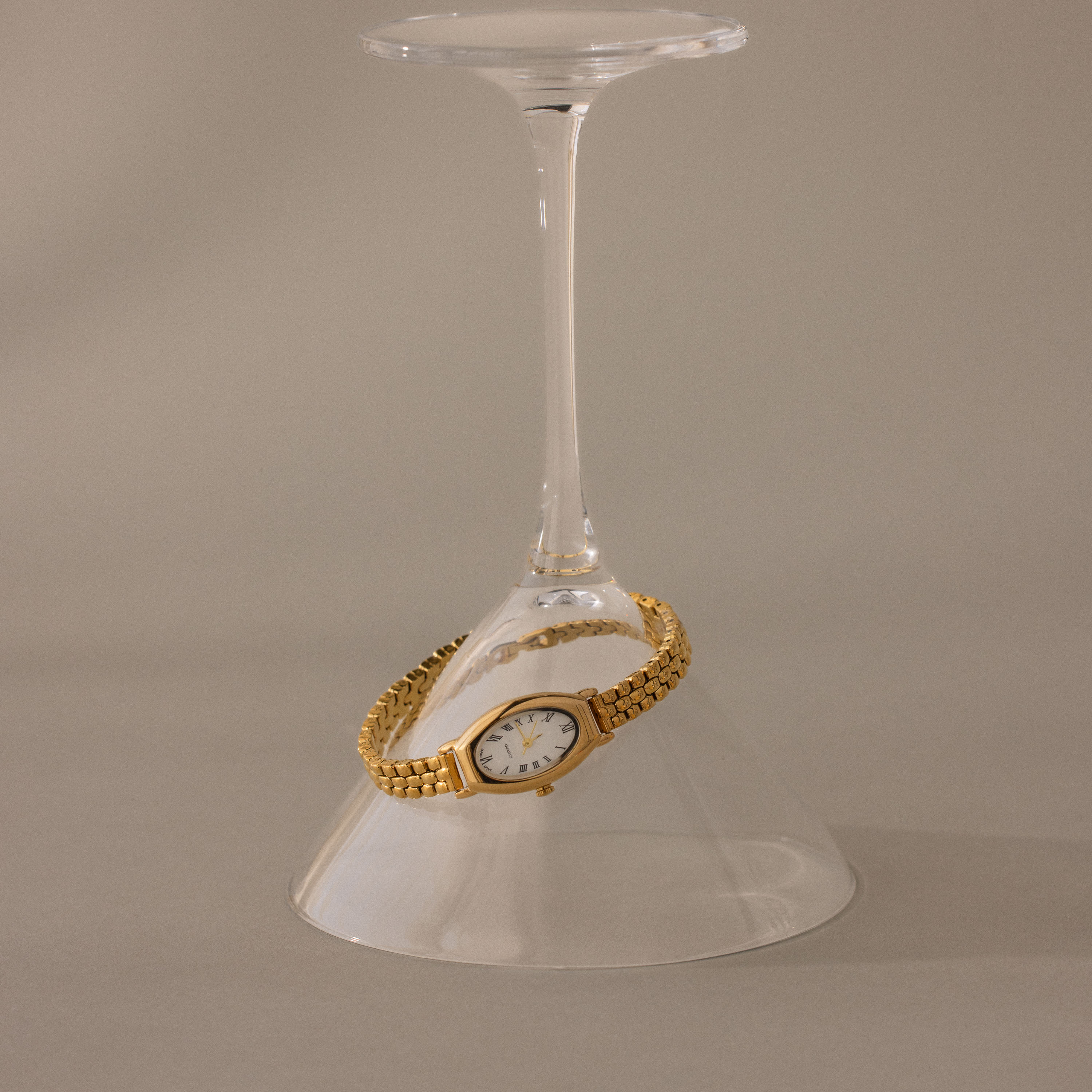 The Grid Chain Timepiece, a vintage-inspired bracelet oval dial watch, is elegantly draped around the stem of an upside-down wine glass on a neutral backdrop.