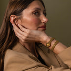 A woman in a tan blazer showcases the Oval Link Timepiece Set—featuring a gold-tone watch and link bracelet—with her hand on her neck, looking to the side.