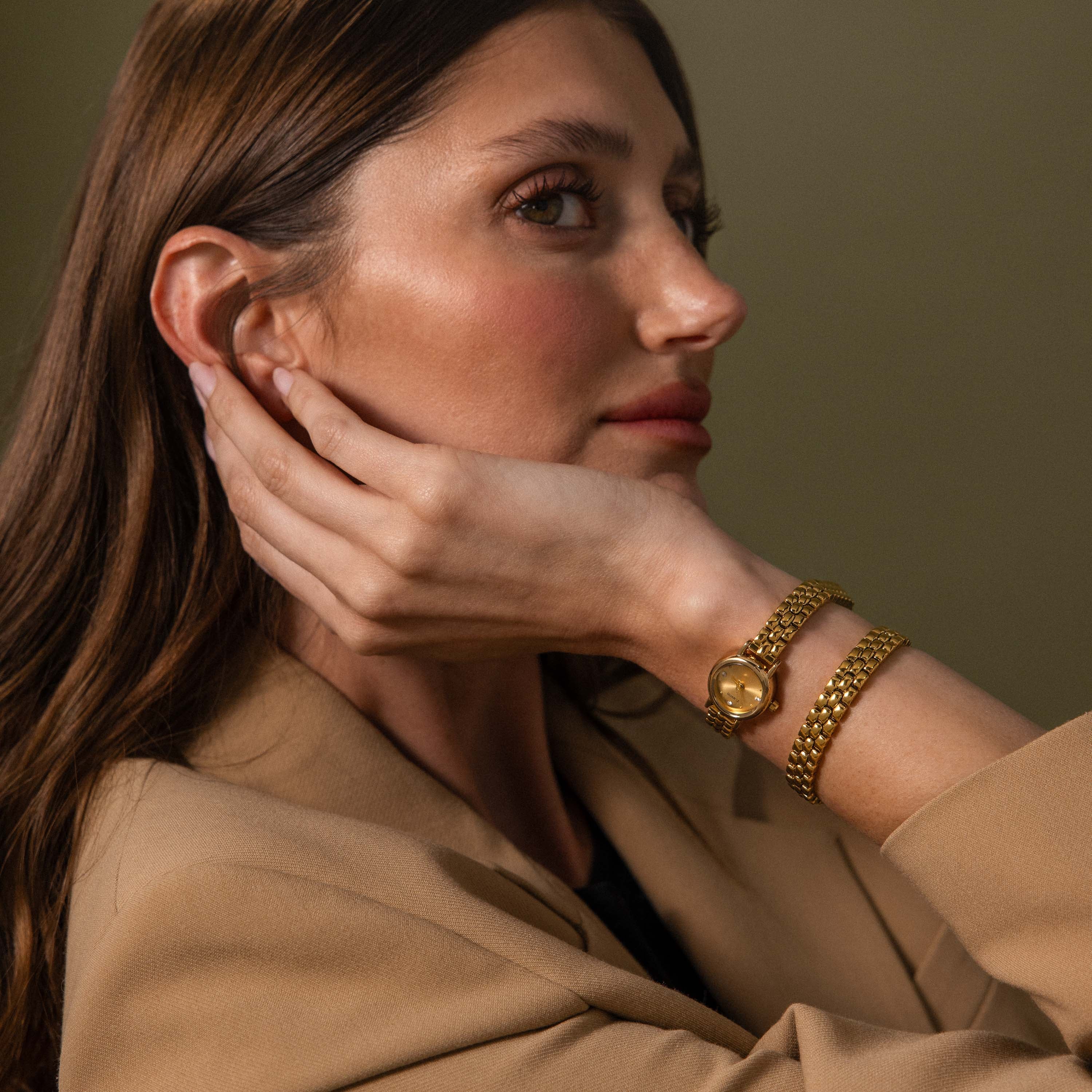 A woman in a tan blazer showcases the Oval Link Timepiece Set—featuring a gold-tone watch and link bracelet—with her hand on her neck, looking to the side.