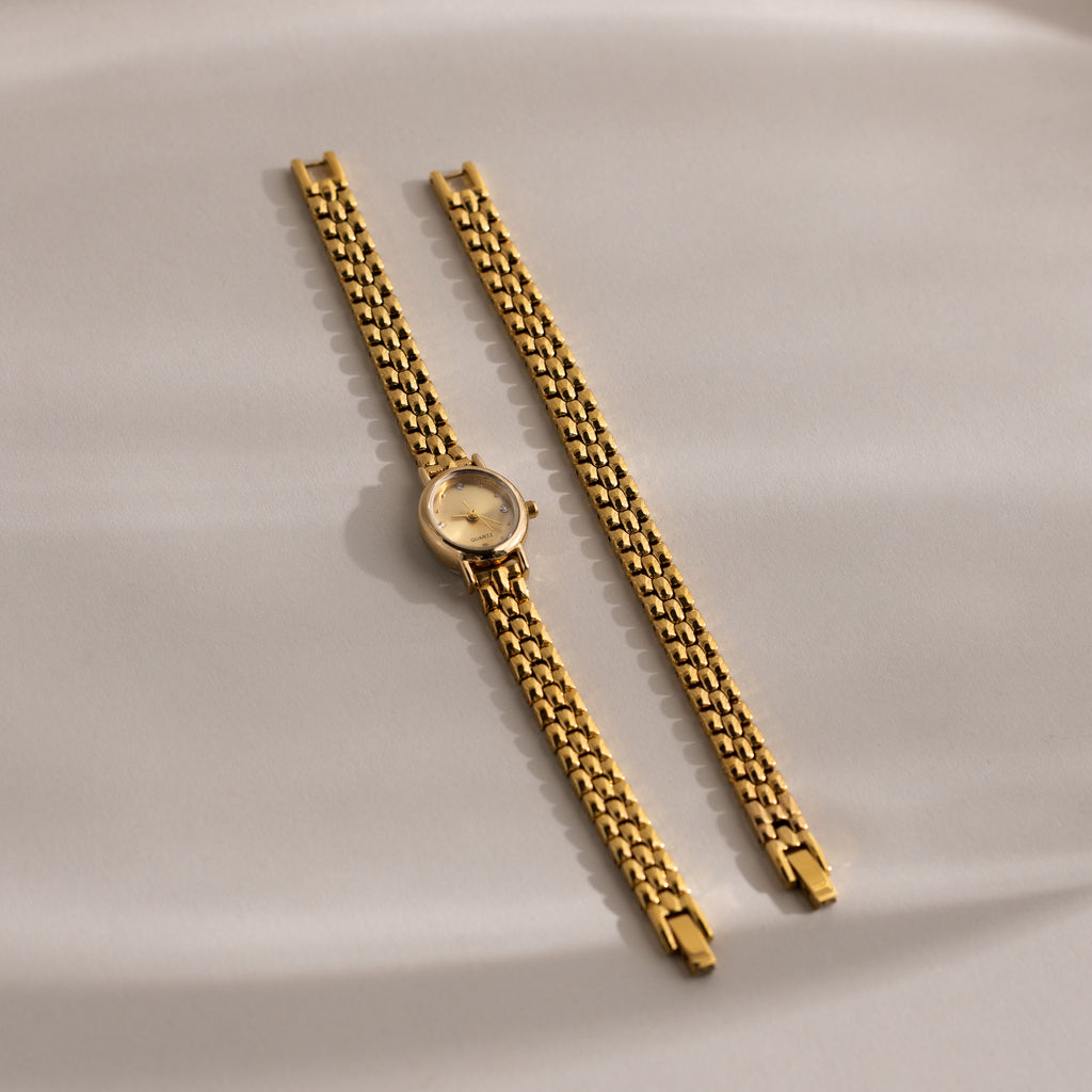 The Oval Link Timepiece Set, featuring a gold-tone watch and its detached link bracelet, is showcased on a smooth beige surface for an elegant look.