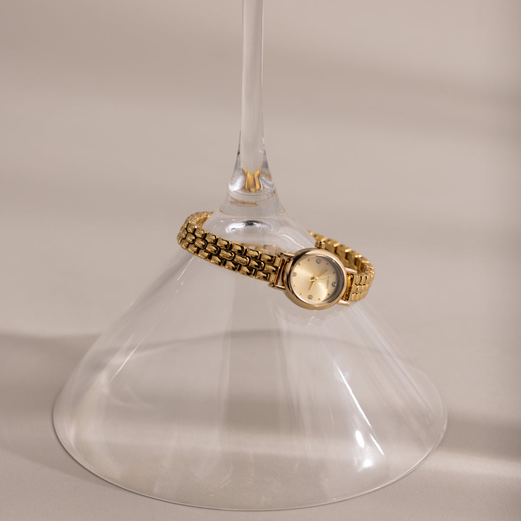 The Oval Link Timepiece Set, featuring a gold-tone watch with a link bracelet, wraps around the stem of an upside-down wine glass on a beige surface.