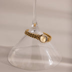 The Oval Link Timepiece Set, featuring a gold-tone watch with a link bracelet, wraps around the stem of an upside-down wine glass on a beige surface.