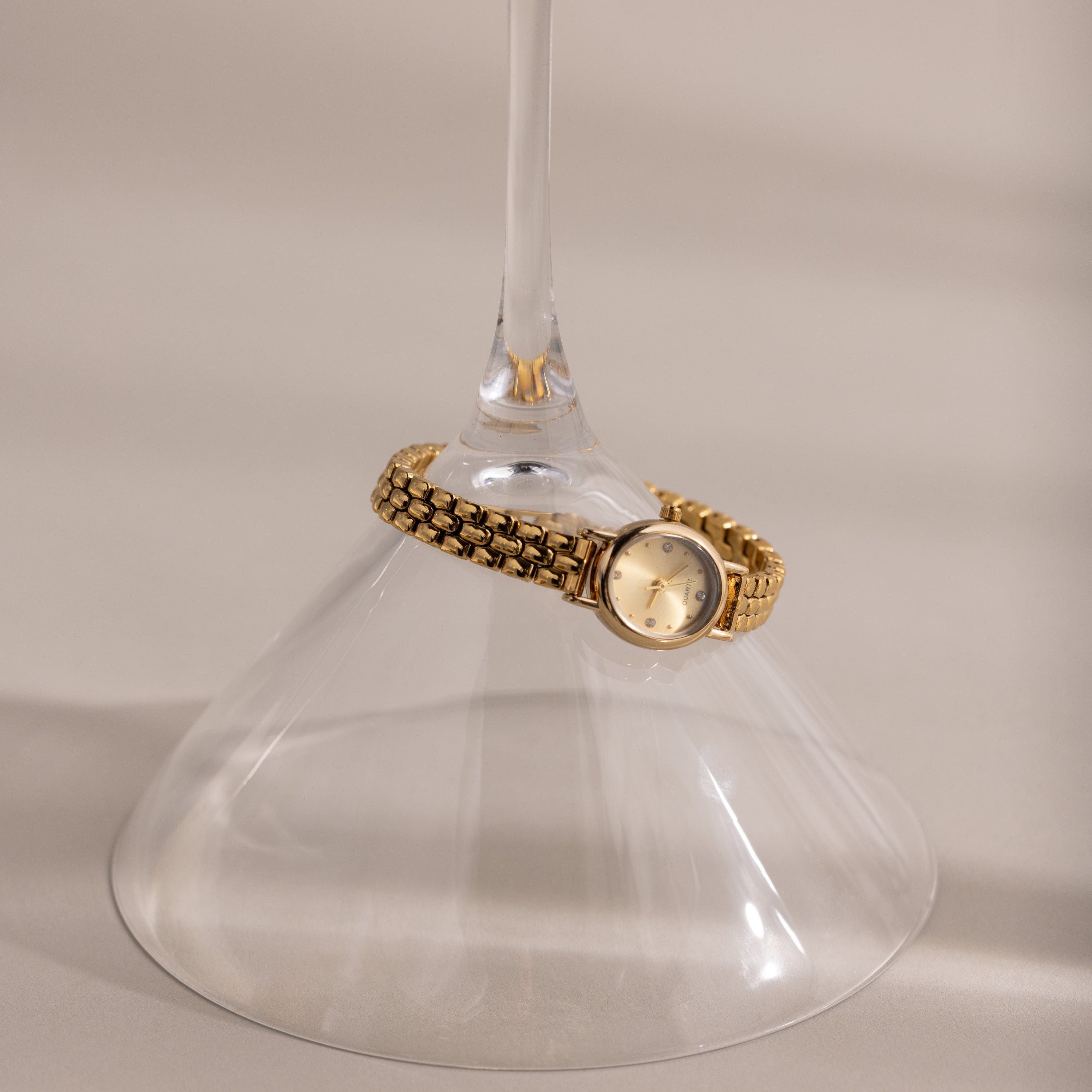 The Oval Link Timepiece Set, featuring a gold-tone watch with a link bracelet, wraps around the stem of an upside-down wine glass on a beige surface.