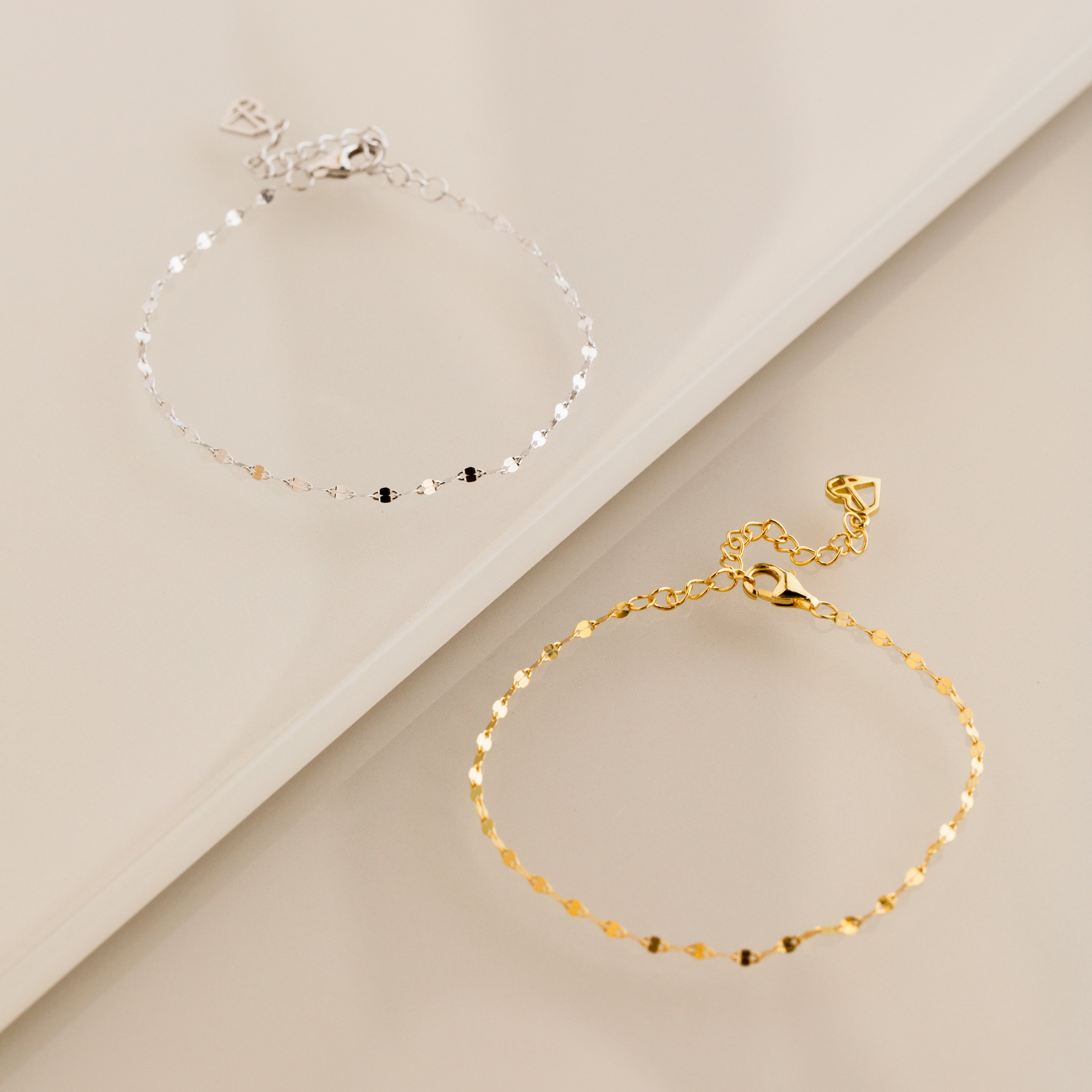 The Josephine Chain Bracelet in gold and a silver anchor link bracelet are displayed on a glossy beige surface.