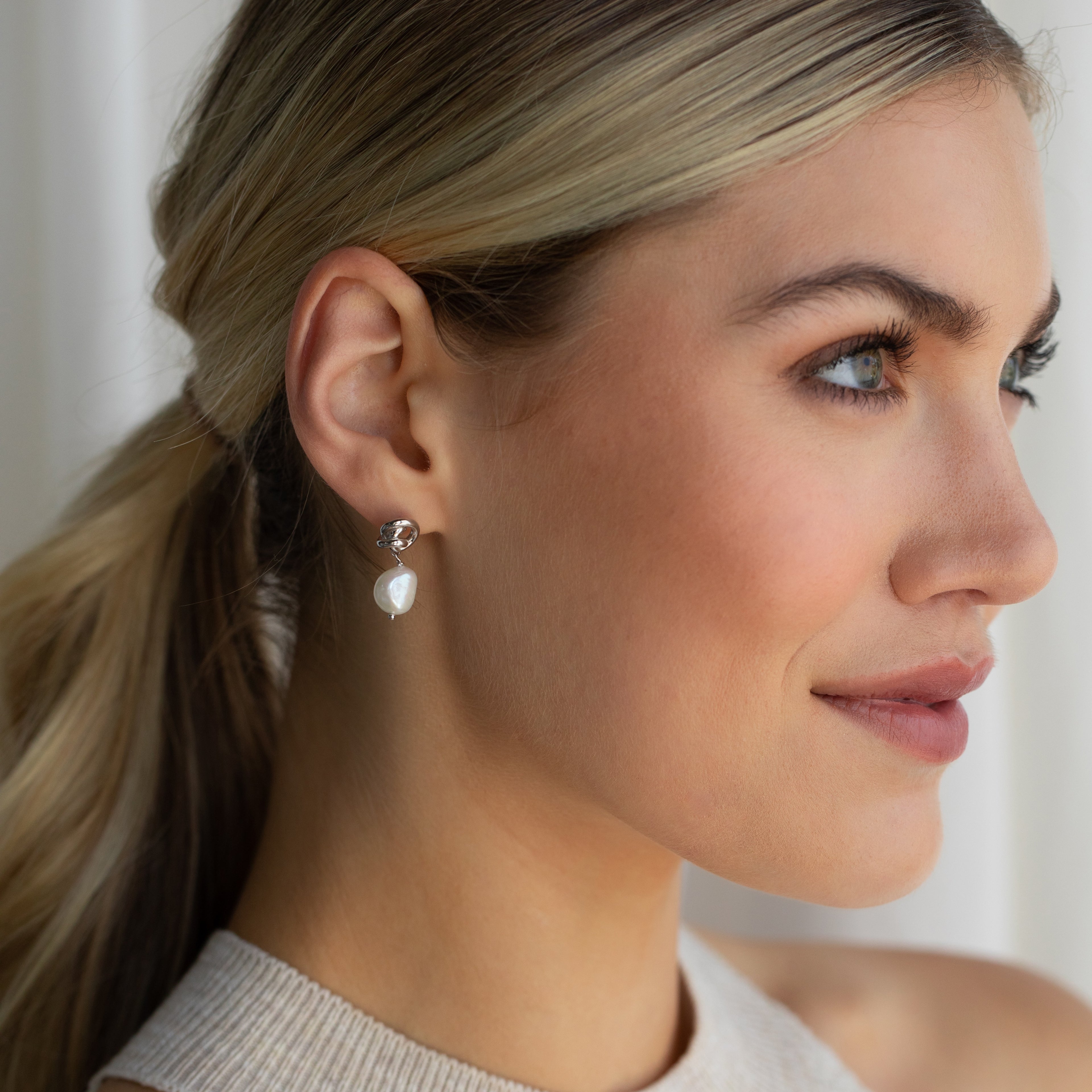 A blonde woman with her hair in a low ponytail smiles softly while wearing Knot Pearl Drop Earrings in Sterling Silver and gazing to the side.