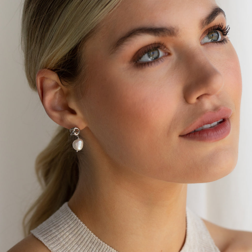 A woman with light eyes and nude lipstick wears Knot Pearl Drop Earrings in Sterling Silver, gazing upward in soft natural light.