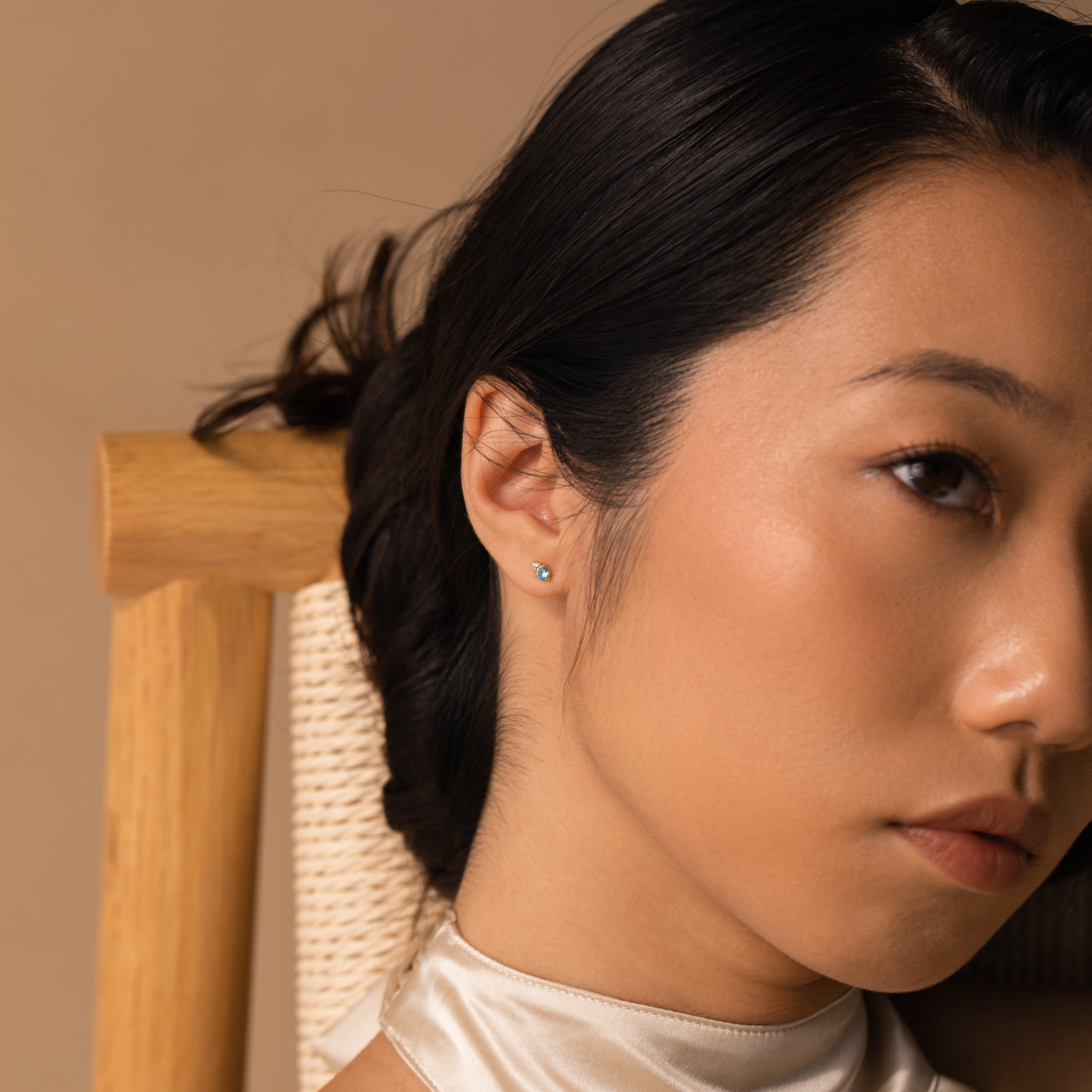 Close-up of a woman with smooth skin, wearing Raindrop Aquamarine Studs, sitting on a wooden chair against a beige background.