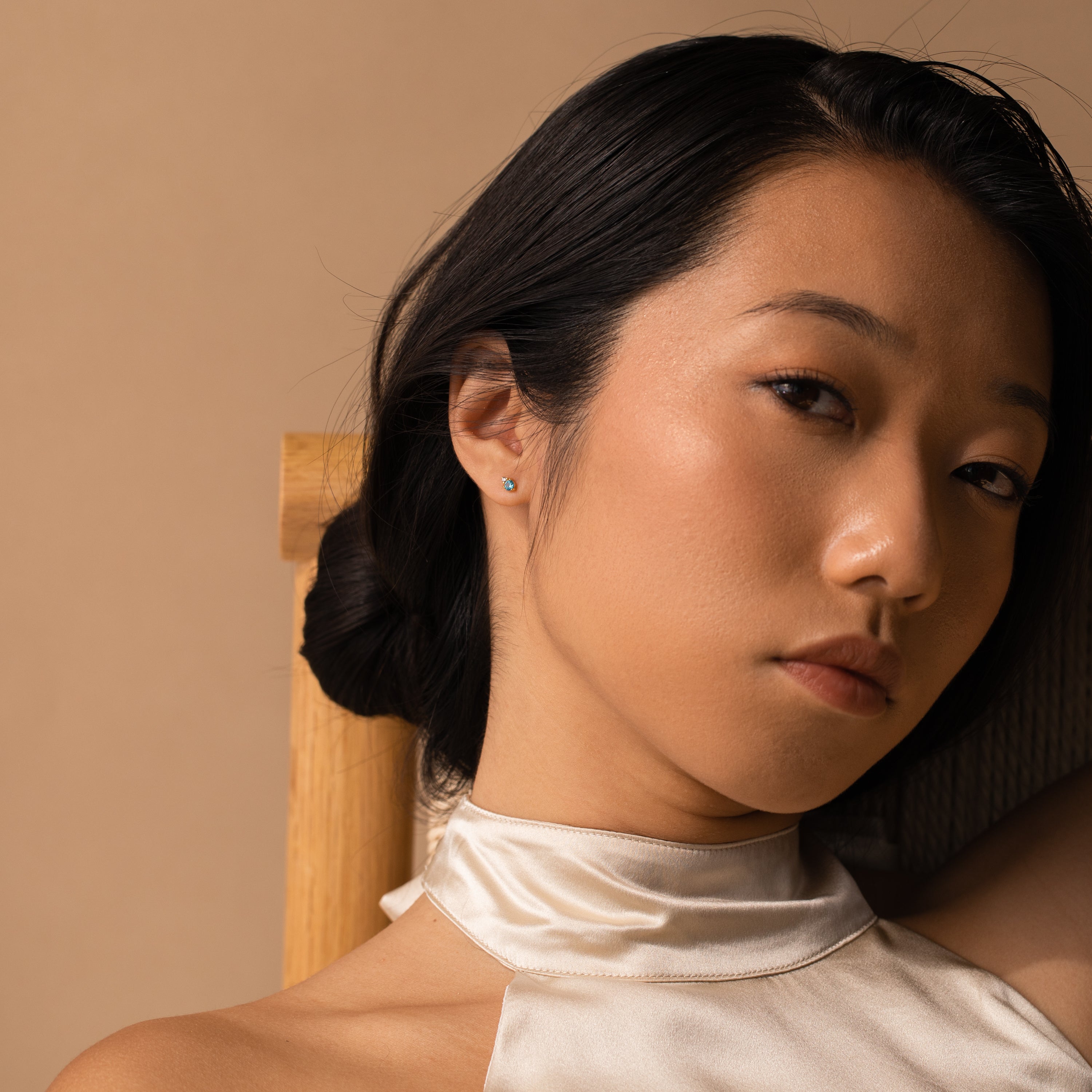 Woman with straight black hair in a silk halter top sits on a wooden chair, gazing softly at the camera, her Raindrop Birthstone Studs adding a touch of subtle elegance.