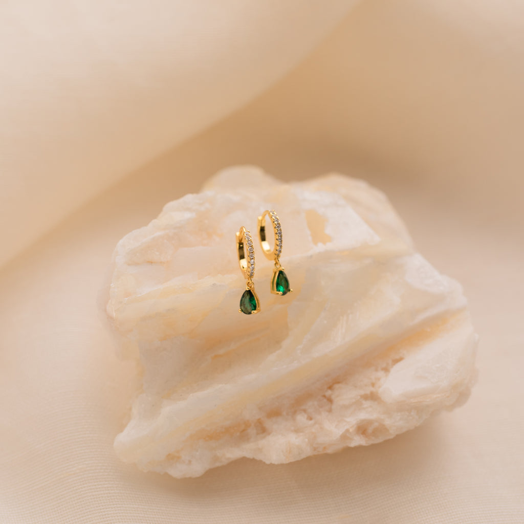 Gold hoop earrings with green birthstone teardrops, shown on a textured white stone background, highlight the elegance of Pave Birthstone Drop Huggies.