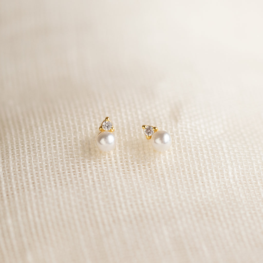 The Raindrop Birthstone Studs are gold earrings with white pearls and small diamonds, shown on a textured cream fabric background.