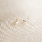 The Raindrop Birthstone Studs are gold earrings with white pearls and small diamonds, shown on a textured cream fabric background.