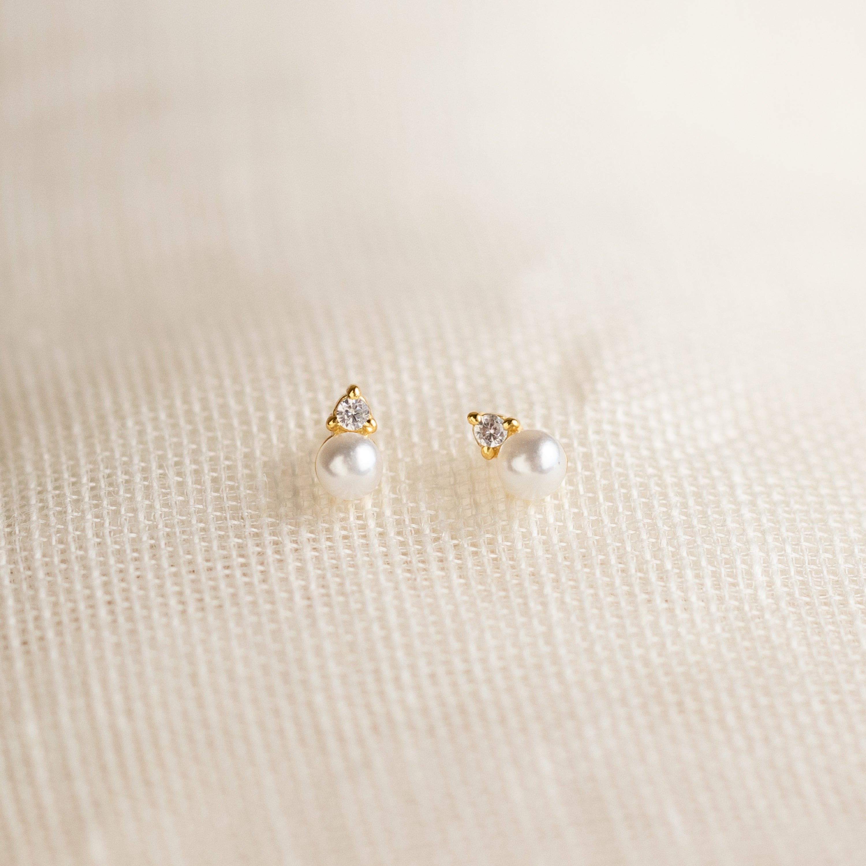 The Raindrop Birthstone Studs are gold earrings with white pearls and small diamonds, shown on a textured cream fabric background.