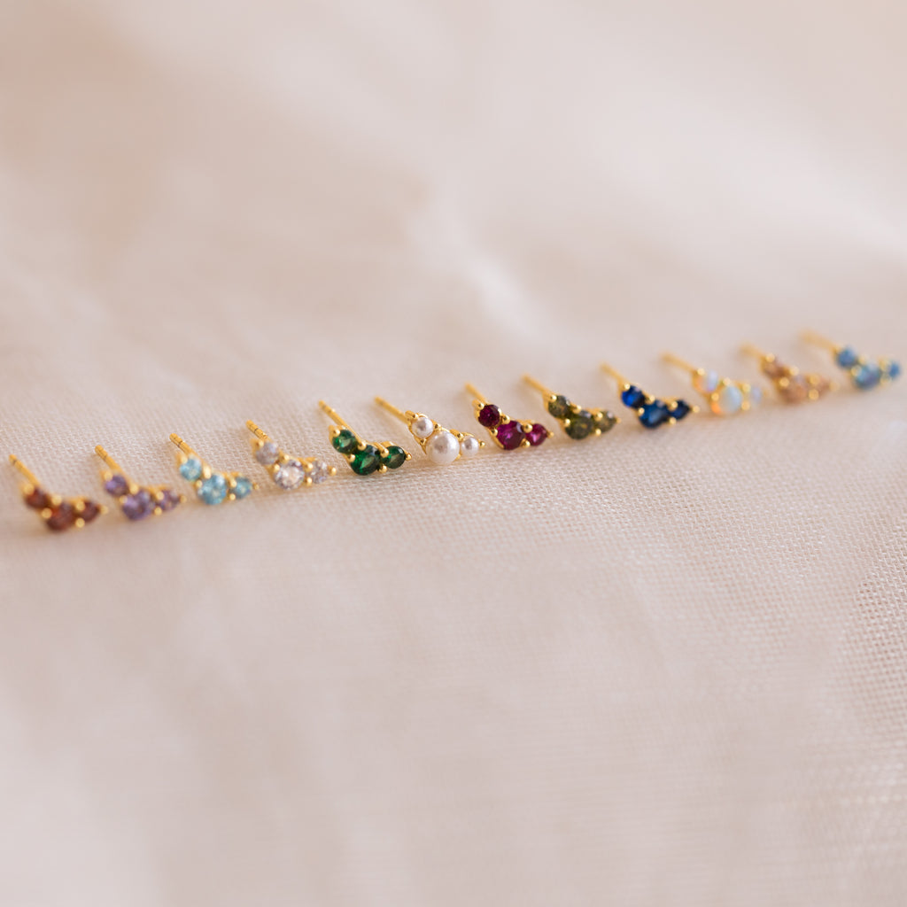 Bella Emerald Studs—small, vibrant green birthstone studs set in gold—are displayed on white fabric, making an ideal personalized jewelry gift for mothers and daughters.