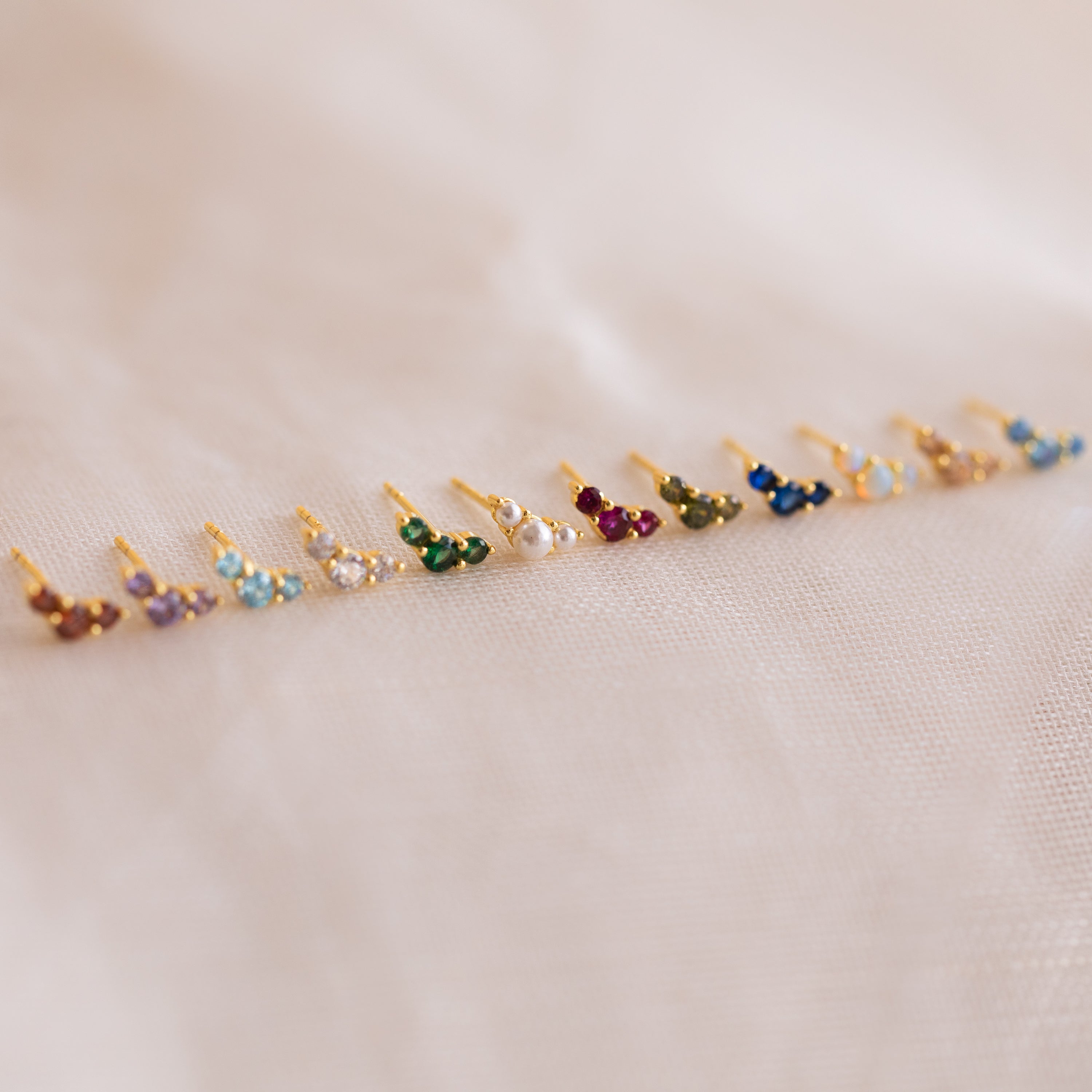 Bella Emerald Studs—small, vibrant green birthstone studs set in gold—are displayed on white fabric, making an ideal personalized jewelry gift for mothers and daughters.