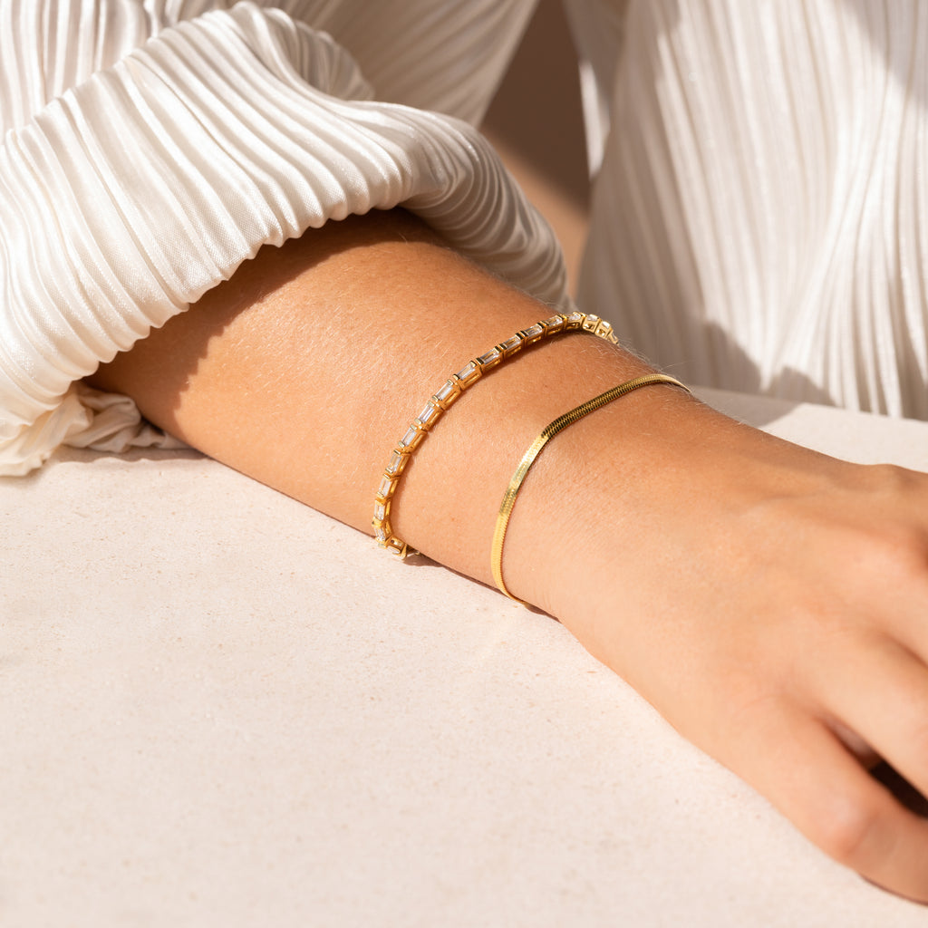A person wearing two gold bracelets, including the Claudia Baguette Tennis Bracelet, on their wrist, paired with a white pleated long-sleeve top.