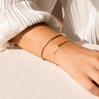 A person wearing two gold bracelets, including the Claudia Baguette Tennis Bracelet, on their wrist, paired with a white pleated long-sleeve top.