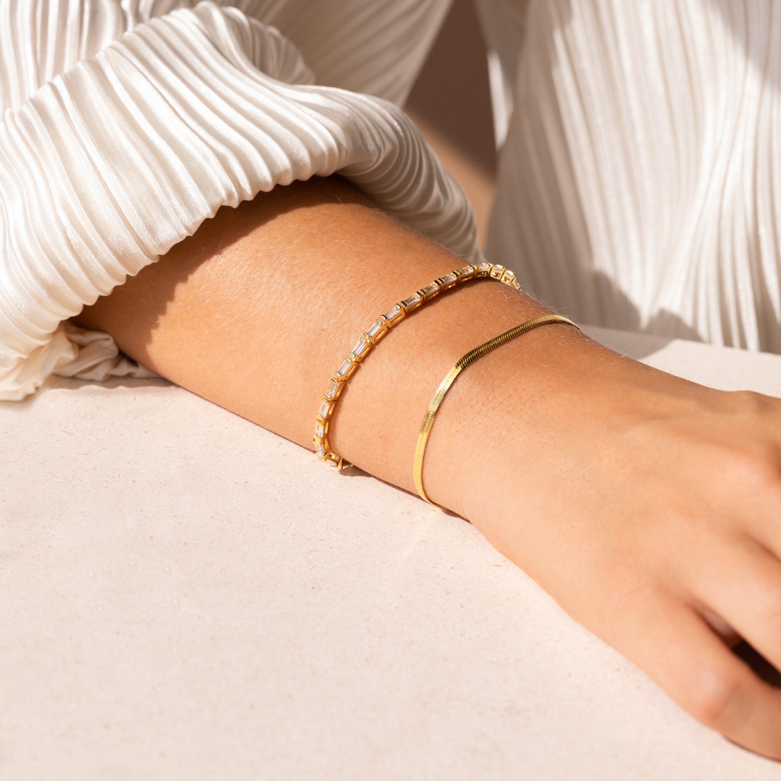 A person wearing two gold bracelets, including the Claudia Baguette Tennis Bracelet, on their wrist, paired with a white pleated long-sleeve top.