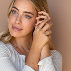 A blonde woman in a white top touches her face, showcasing the elegant Thin Stacking Rings Set in 18K Gold—an ideal Christmas gift—as she looks off camera.