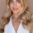 Smiling woman with long blonde hair wears a white top and the Initial Pendant Necklace, standing against a beige background.