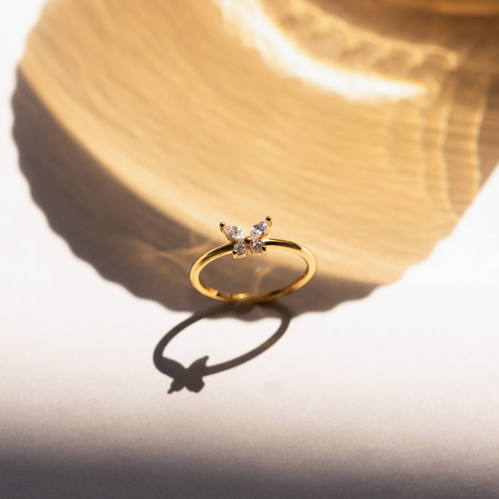 The Mariposa Diamond Ring, featuring a gold butterfly design, casts an elegant shadow on a light surface under bright sunlight.