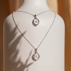 Two Initial Pendant Necklaces in Sterling Silver with round M and K pendants are displayed on a white stand—perfect for adding a vintage-inspired touch to any look.