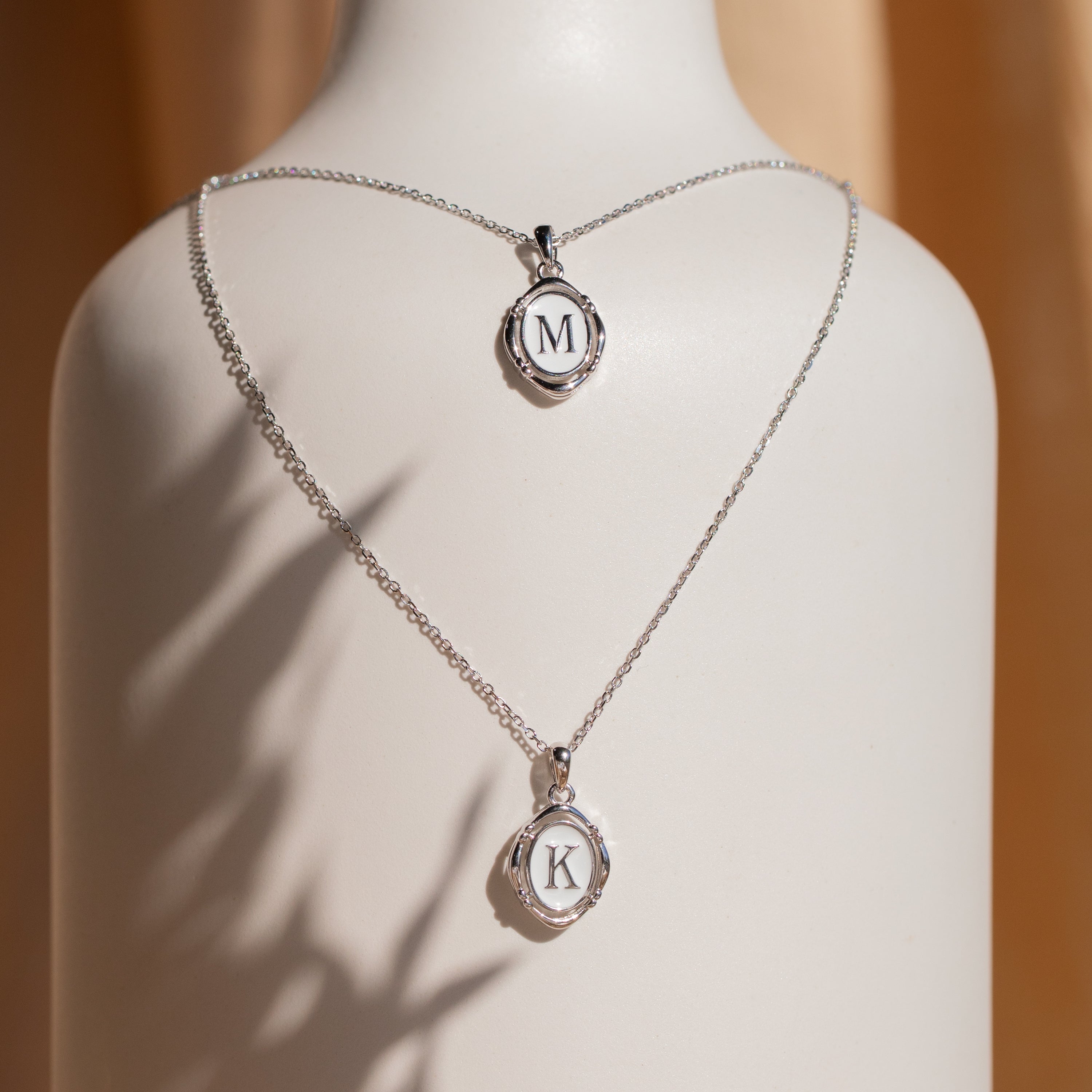 Two Initial Pendant Necklaces in Sterling Silver with round M and K pendants are displayed on a white stand—perfect for adding a vintage-inspired touch to any look.
