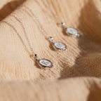 The Initial Pendant Necklace in Sterling Silver rests on textured beige fabric, softly illuminated by natural sunlight, showcasing its vintage-inspired round pendant style.