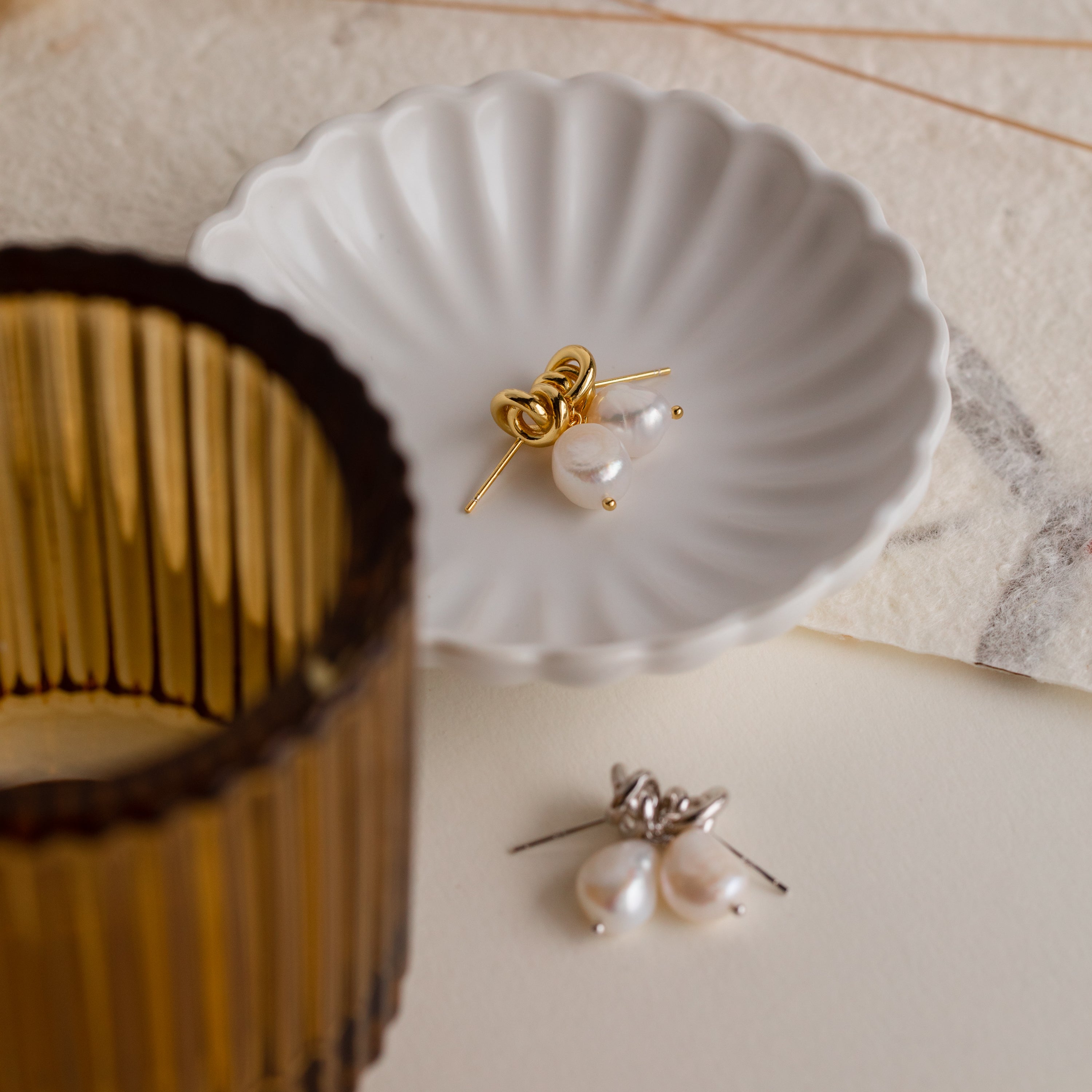 The Knot Pearl Drop Earrings in Sterling Silver are displayed in a white dish beside a ribbed amber glass—an elegant option for those who appreciate minimalist jewelry.