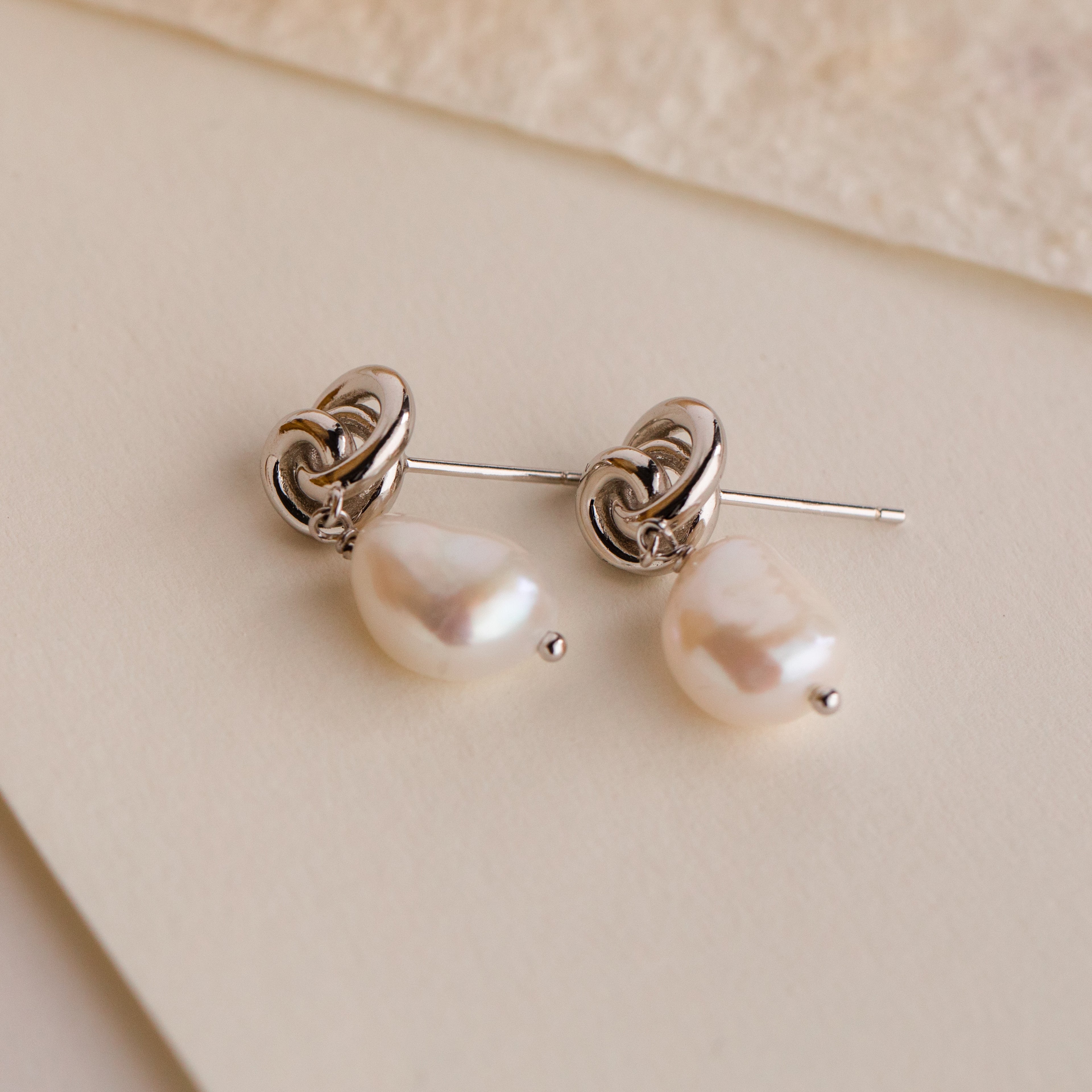 Knot Pearl Drop Earrings featuring dangling white pearls, displayed on cream-colored textured paper—perfect minimalist earrings for an elegant touch.