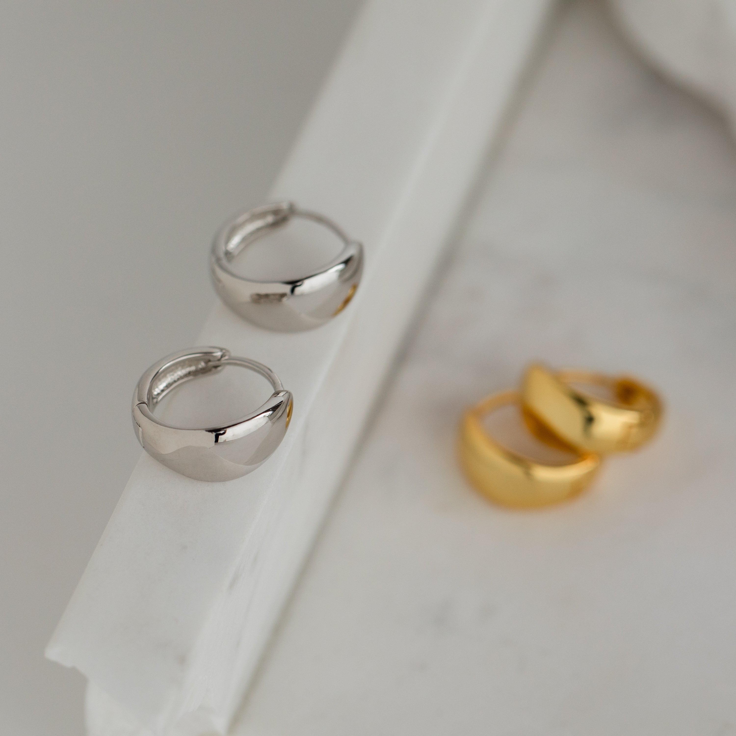 The Olivia Hoops in Sterling Silver—chunky, modern earrings—are displayed on a white marble surface, perfect for adding a contemporary touch to any outfit.