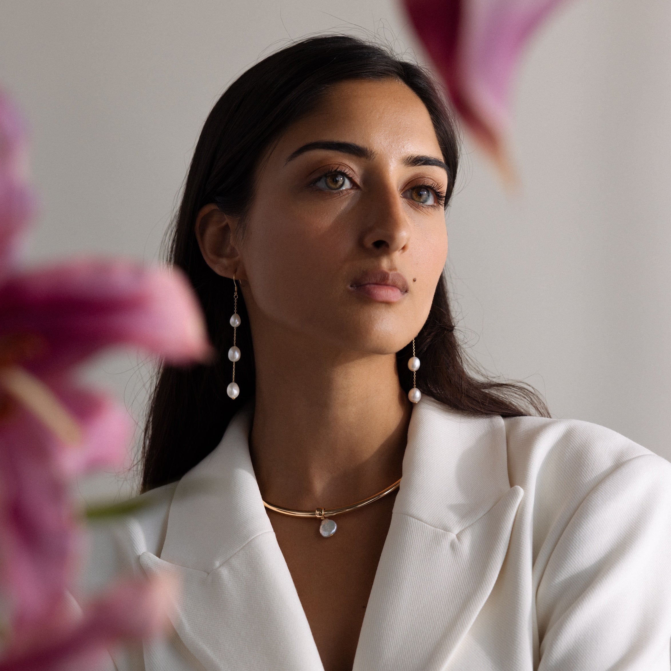 Woman in a white blazer wears baroque pearl earrings and a Pearl Cuff Choker Necklace in 18K Gold, with pink flowers in the foreground.