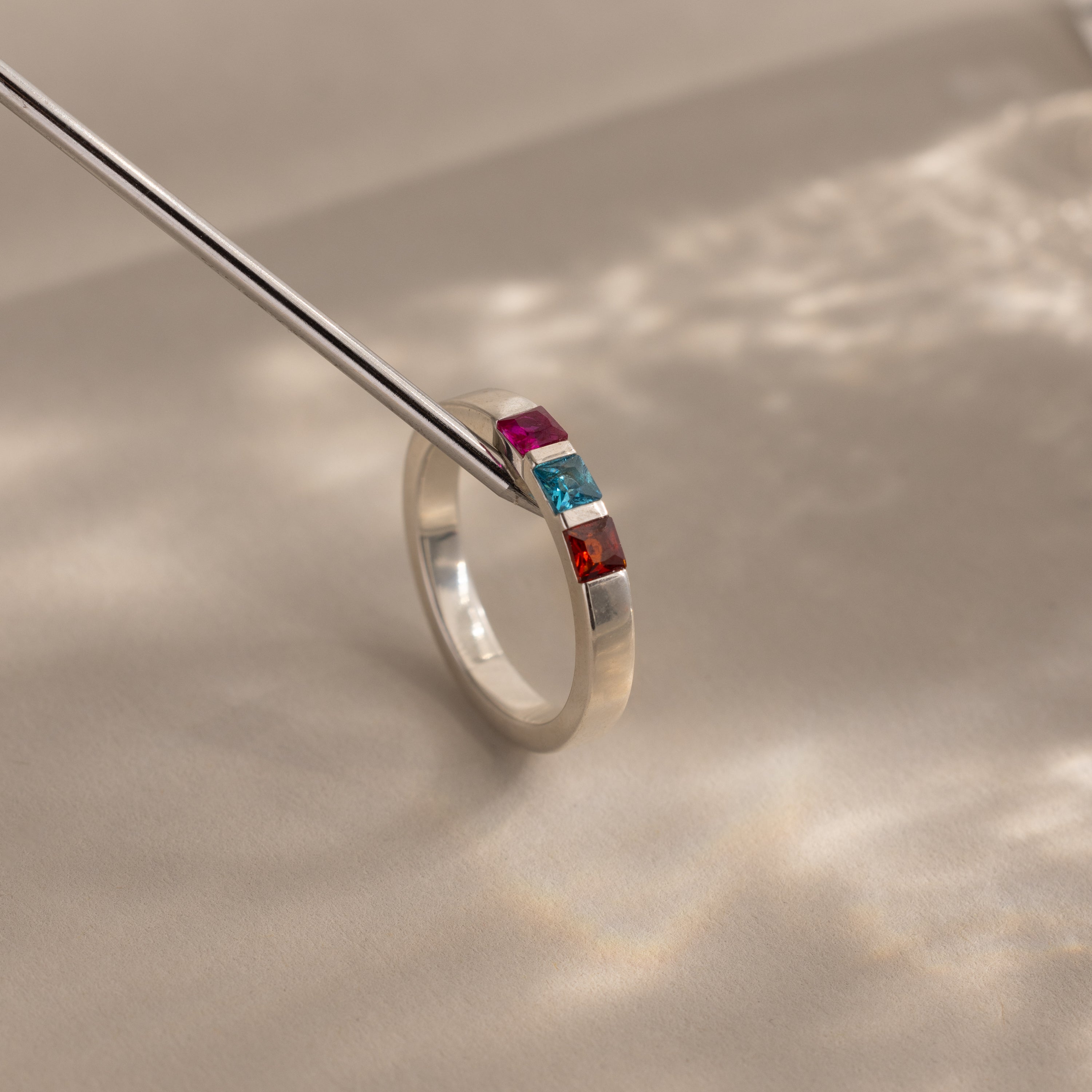 The Multiple Birthstone Band in Sterling Silver features pink, blue, and red rectangular gemstones on a neutral background—ideal as a personalized mothers ring.
