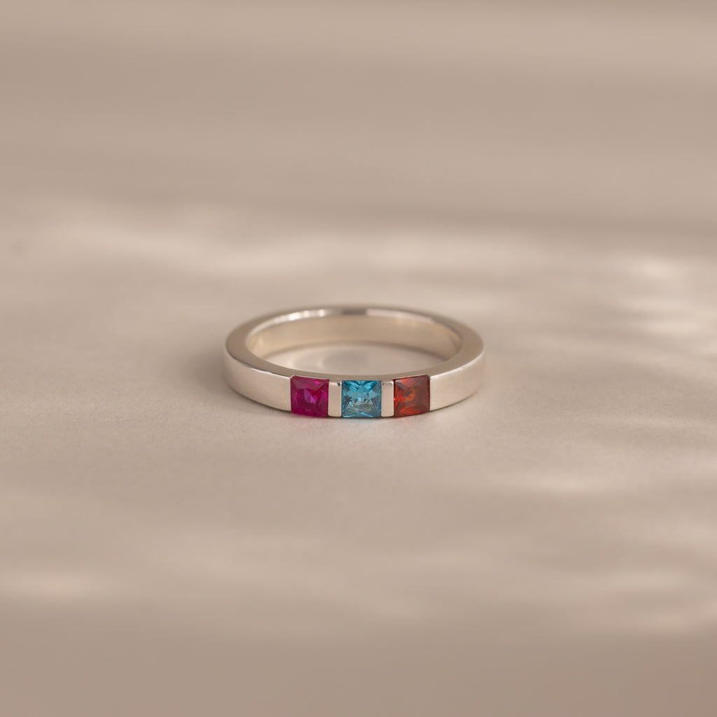 The Multiple Birthstone Band features three square gemstones—pink, blue, and orange—set in silver. This personalized jewelry piece makes a unique birthstone ring, displayed on a neutral background.