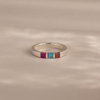 The Multiple Birthstone Band features three square gemstones—pink, blue, and orange—set in silver. This personalized jewelry piece makes a unique birthstone ring, displayed on a neutral background.