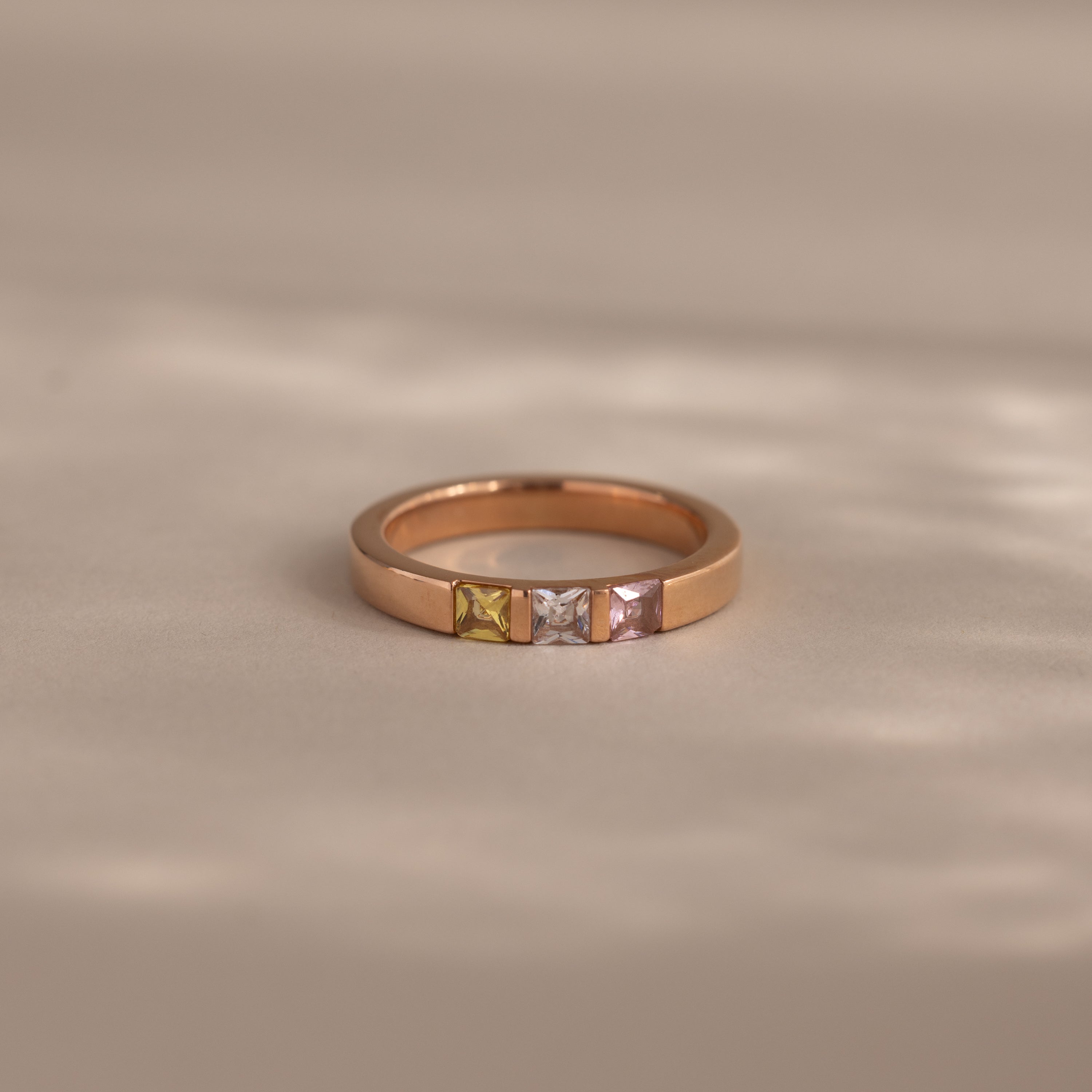 The Multiple Birthstone Band in Rose Gold features four square gemstones in yellow, green, white, and pink on a beige surface—a beautiful personalized jewelry piece and perfect mothers gift.