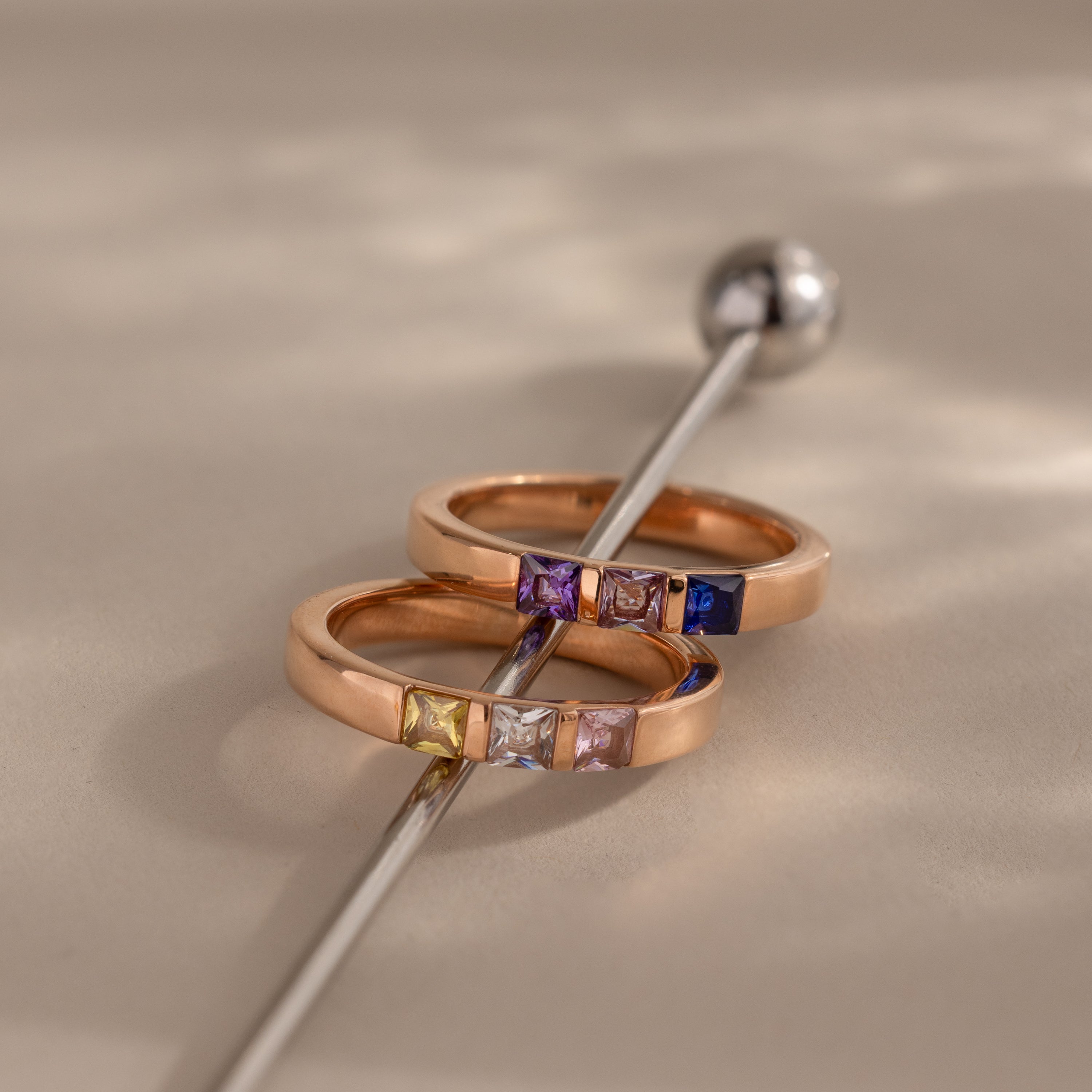 Two Multiple Birthstone Bands in rose gold, set with vibrant square gemstones, lie beside a silver rod on a beige surface—a beautiful Mother’s gift and personalized jewelry piece.
