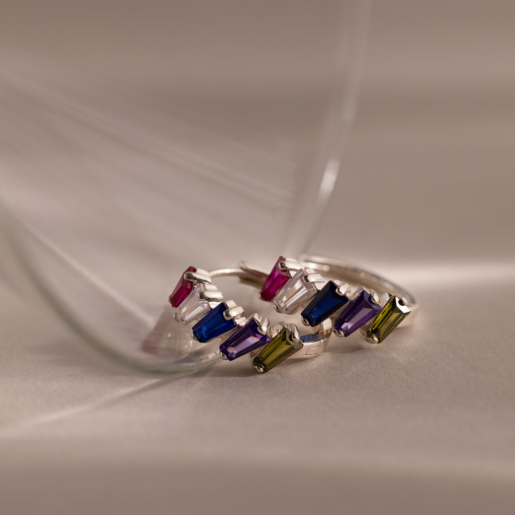 Three Multiple Tapered Birthstone Huggies in Sterling Silver, each set with colorful gemstones, displayed on a soft beige surface with gentle glass reflections.