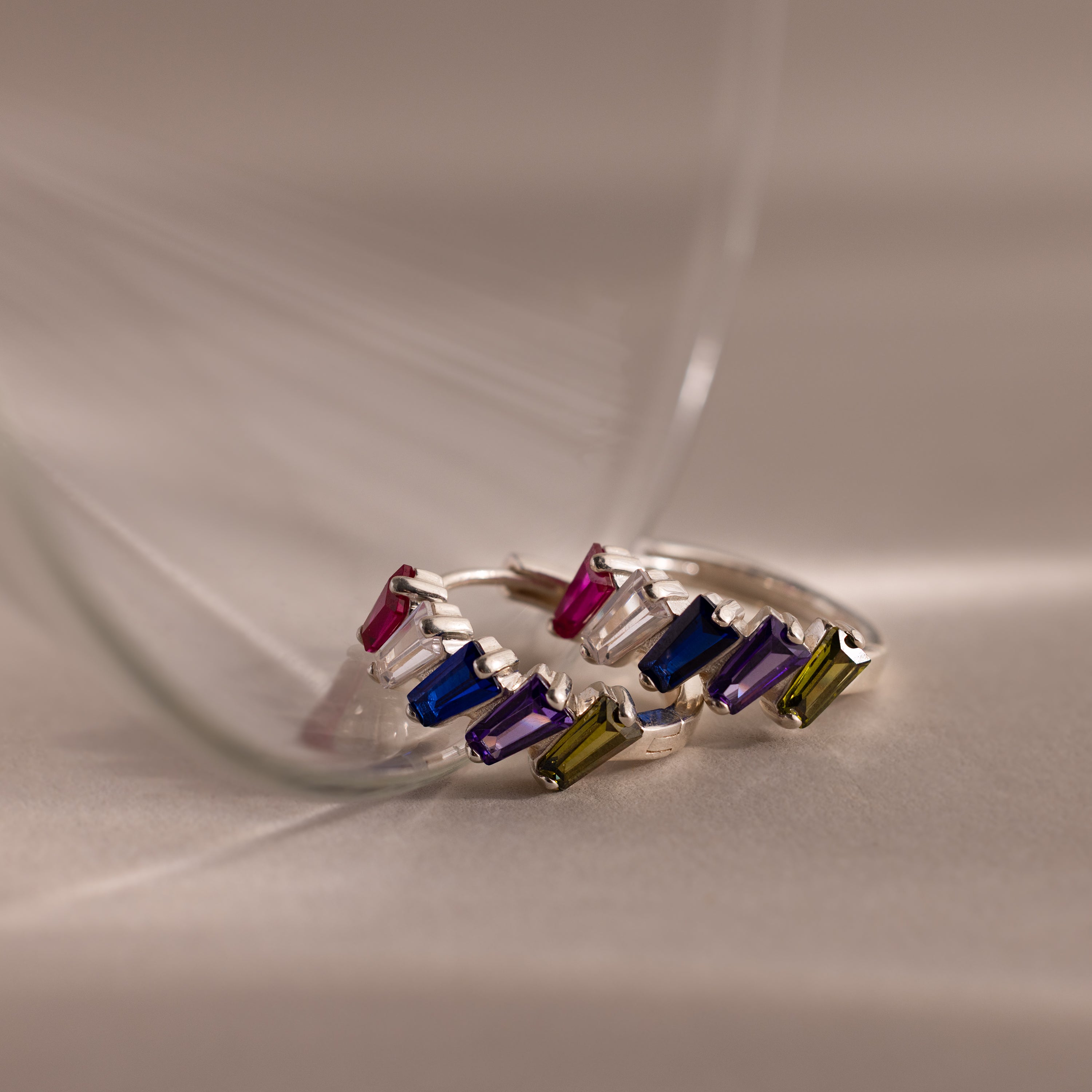 Three Multiple Tapered Birthstone Huggies in Sterling Silver, each set with colorful gemstones, displayed on a soft beige surface with gentle glass reflections.