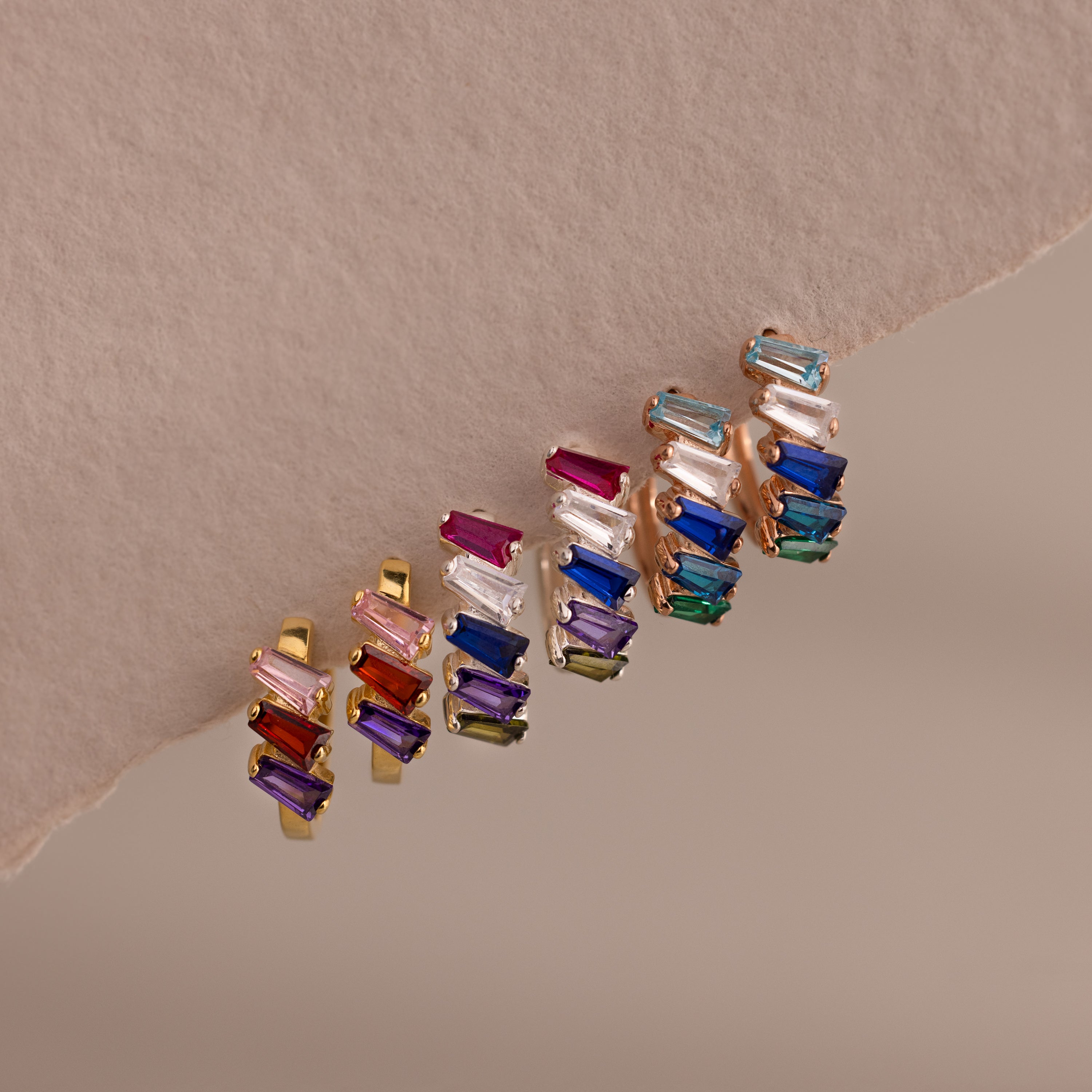 Five pairs of Multiple Tapered Birthstone Huggies with colorful rectangular gemstones are arranged on a textured beige surface, showcasing bold modern statement jewelry.