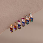Five pairs of rose gold hoop earrings, featuring Multiple Tapered Birthstone Huggies with colorful gemstones, are arranged in a row on a beige background.