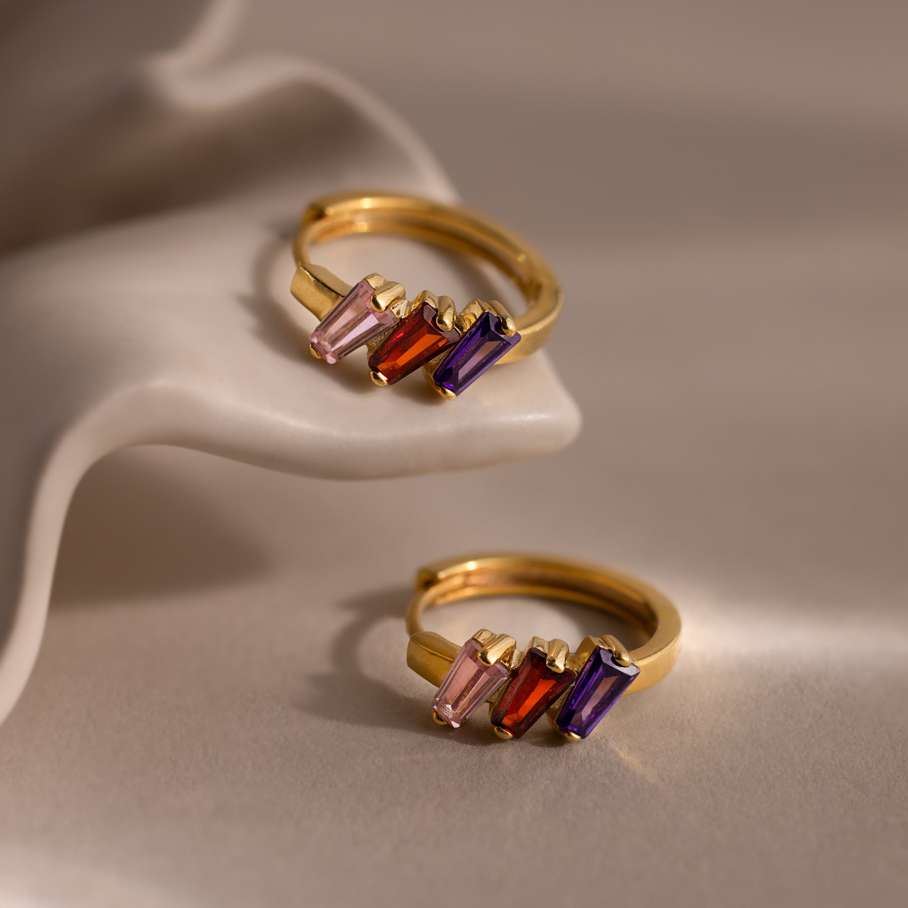 Multiple Tapered Birthstone Huggies in 18K Gold with pink, red, and purple rectangular stones displayed on a white sculpted surface—ideal for fans of modern statement jewelry or unique Art Deco-inspired earrings.